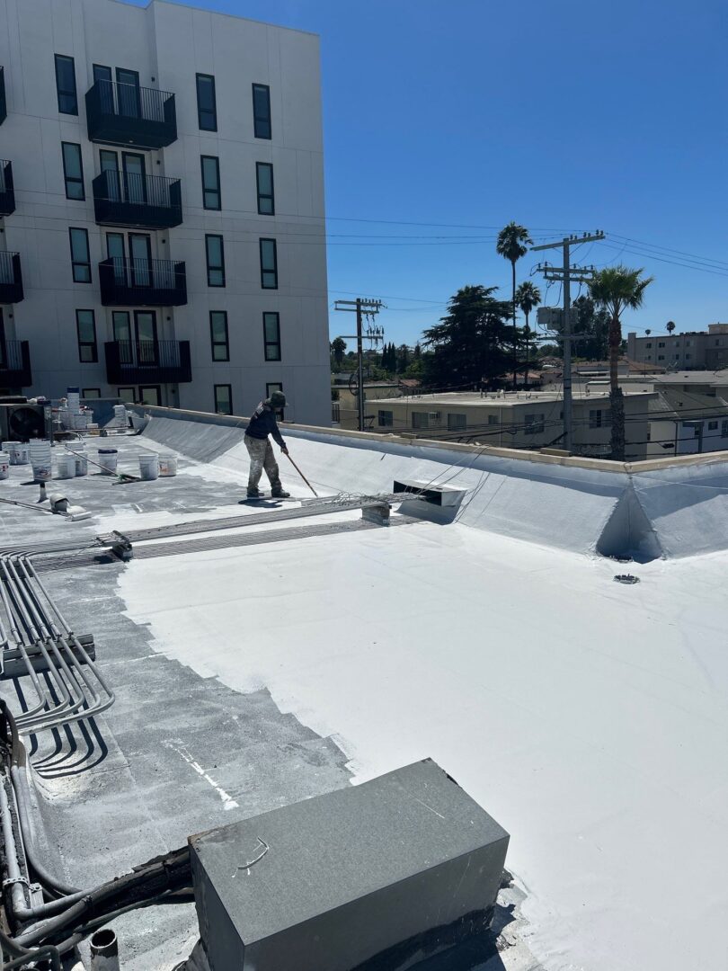 A worker applying white coating on a flat rooftop under clear skies.