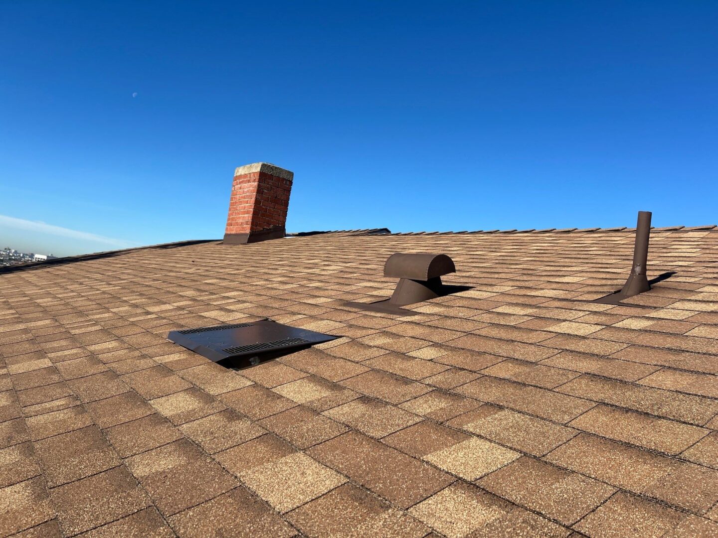 A shingled rooftop with a chimney and vents under a clear blue sky.