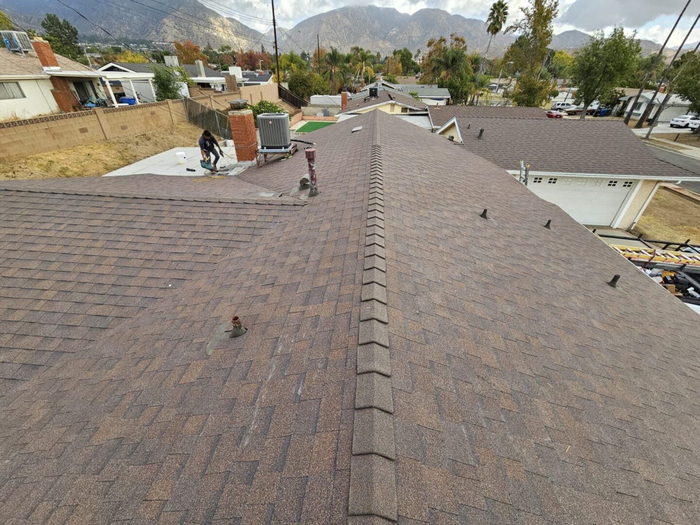 A long, brown shingled roof with a chimney and vents under a cloudy sky.