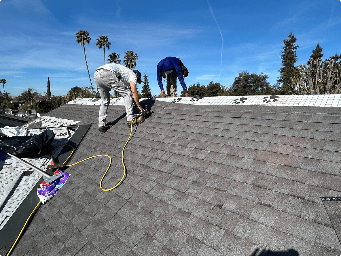 Two workers installing roofing shingles on a sunny day.
